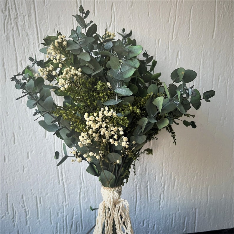 Pode incluir: Um buqu&ecirc; de flores e folhagens secas com folhas de eucalipto verde-mate, pequenos cachos de gips&oacute;fila branca e folhagem de preenchimento verde texturizada. Os caules s&atilde;o reunidos e amarrados com um cord&atilde;o de macram&eacute; na cor creme.
