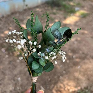 Pode incluir: Um pequeno arranjo floral com folhas de eucalipto verde-escuras, delicadas flores brancas de gips&oacute;fila e ramos verdes finos. Os caules est&atilde;o agrupados e presos com um cordel castanho-claro na base do ramo.