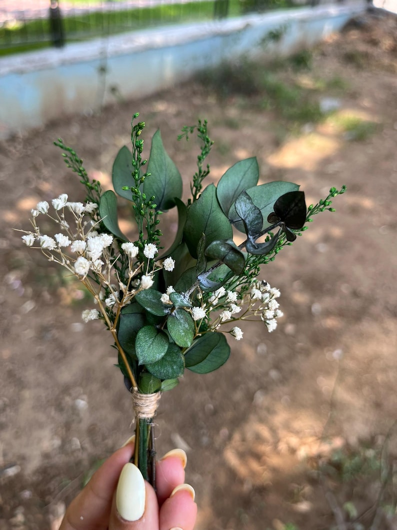 Pode incluir: Um pequeno arranjo floral com folhas de eucalipto verde-escuras, delicadas flores brancas de gips&oacute;fila e ramos verdes finos. Os caules est&atilde;o agrupados e presos com um cordel castanho-claro na base do ramo.