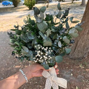 Pode incluir: Um buqu&ecirc; de folhas de eucalipto secas e pequenas flores brancas de gips&oacute;fila. Os caules est&atilde;o reunidos e envolvidos por uma fita larga de renda cor de creme com trama aberta, atada num la&ccedil;o na base do arranjo floral.