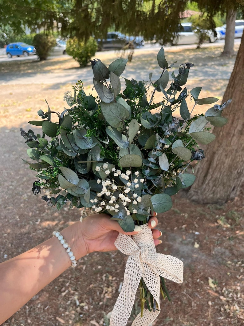 Pode incluir: Um buqu&ecirc; de folhas de eucalipto secas e pequenas flores brancas de gips&oacute;fila. Os caules est&atilde;o reunidos e envolvidos por uma fita larga de renda cor de creme com trama aberta, atada num la&ccedil;o na base do arranjo floral.