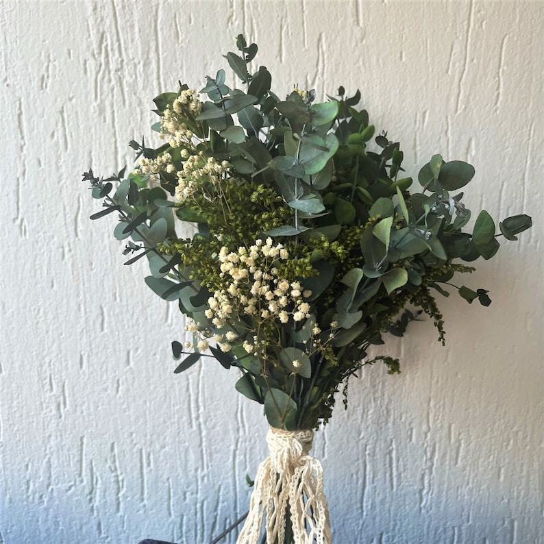 Pode incluir: Um buqu&ecirc; de ramos de eucalipto secos com folhas arredondadas verde-azuladas, intercaladas com cachos de pequenas flores brancas de gips&oacute;fila e folhagem verde texturizada. Os caules est&atilde;o reunidos e atados com uma fita de renda cor de creme.