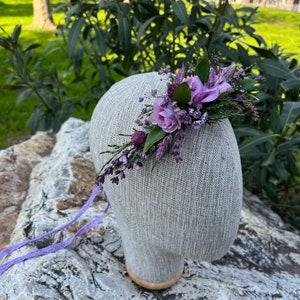 May include: A floral headpiece featuring lavender and purple flowers, greenery, and sprigs. The arrangement is attached to a neutral-colored head form. Lavender ribbons extend from the base of the head form.