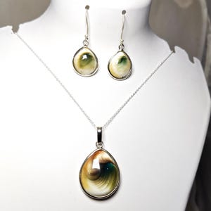 Op de afbeelding: Een sieradenset bestaande uit een zilveren ketting met een druppelvormige hanger en bijpassende oorbellen. Elk sieraad is voorzien van een gepolijste schelpinleg met wervelende patronen in crème, bruin en donkergroen, gezet in zilverkleurig metaal.