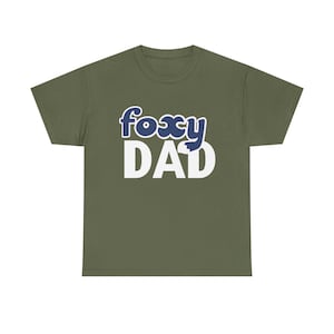 Könnte beinhalten: Olivgrünes T-Shirt mit dem Schriftzug "foxy DAD" in weißen Blockbuchstaben. Das Wort "foxy" ist in einer blauen Bubble-Schrift mit weißer Umrandung gehalten. Ein lässiges T-Shirt.
