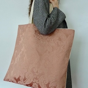 Può includere: Una grande borsa tote con un motivo damascato rosa e manici color crema. La borsa è realizzata in un tessuto morbido e testurizzato.