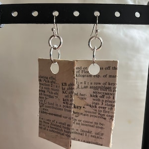 Puede incluir: Un par de pendientes de plata con un diseño circular. Cada pendiente está unido a un pequeño trozo de papel con texto de un libro. El texto en el pendiente izquierdo dice "key" y el texto en el pendiente derecho dice "kick off vb 1 with a place ings 3 slang".