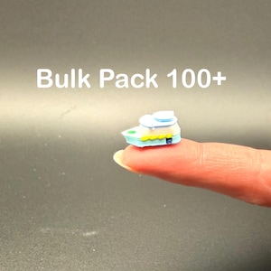 May include: A miniature boat, primarily light blue, white, and yellow, is held on a fingertip. The boat has a small cabin and a yellow trim. The text "Bulk Pack 100+" is displayed in white letters above the boat.