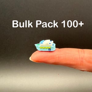 May include: A miniature blue and white boat-shaped charm with yellow accents. The charm is held on a finger. The text "Bulk Pack 100+" is displayed in white letters above the charm.