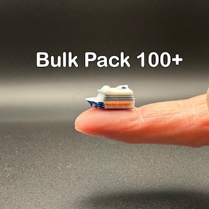 May include: A miniature cruise ship figurine, white with blue and orange accents, is held on a fingertip. The text "Bulk Pack 100+" is displayed above the ship. The ship has a detailed design, including windows and a deck.