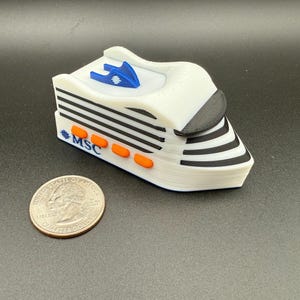 May include: A white cruise ship model with black horizontal stripes, orange rectangular accents, and a blue fin-shaped detail on top. The side of the ship features the blue text MSC. A United States quarter coin is placed next to the ship for scale.