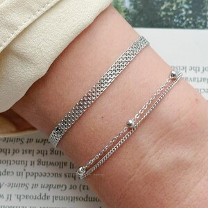 May include: Two silver bracelets. One is a mesh design, and the other is a delicate chain with small silver beads. The bracelets are worn on a wrist, with a cream-colored sleeve in the background. The bracelets are simple and elegant.