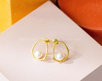 May include: A pair of gold earrings with a pearl in each. The earrings are in the shape of an oval with a cutout in the middle. The earrings are on a pink surface.