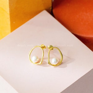 May include: A pair of gold earrings with a pearl in each. The earrings are in the shape of an oval with a cutout in the middle. The earrings are on a pink surface.