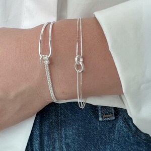 May include: Two silver bracelets are displayed on a wrist. One bracelet has a chain design with a loop and a dangling chain. The other bracelet has a loop design with a chain. The background is a white shirt and blue jeans.