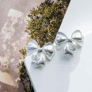 May include: A pair of silver bow-shaped earrings. The earrings are on a white surface.