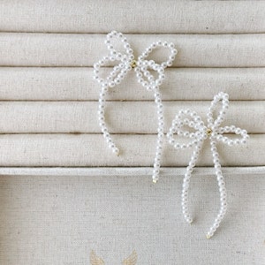 May include: Two white pearl bow hair clips displayed on a white fabric background. The bows are crafted from small white pearls and tied in a simple bow shape. These hair clips are a simple and elegant accessory for any occasion.