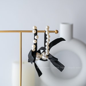 May include: A pair of elegant earrings featuring faux pearls and black satin ribbons. The earrings are displayed on a gold stand against a soft, neutral background. The earrings have a classic design with a modern twist.