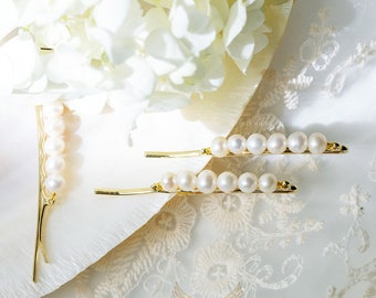 Puede incluir: Tres pinzas para el pelo doradas con una fila de perlas blancas en cada pinza. Las pinzas están sobre una superficie blanca con un diseño floral blanco.