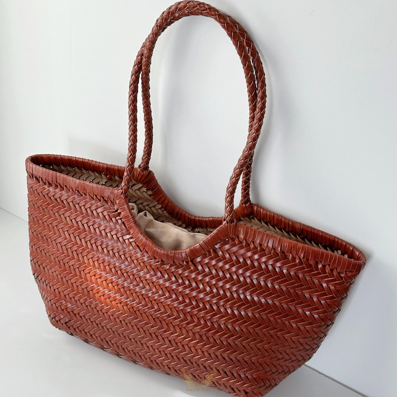 May include: A woven brown tote bag with braided handles. The bag has a wide opening and a light-colored interior. The weave pattern is visible throughout the bag's exterior, and the handles are thick and sturdy.