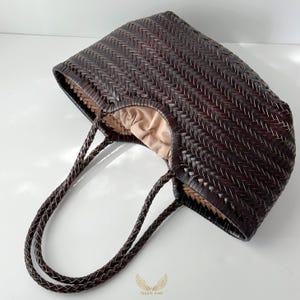 May include: A dark brown woven tote bag with braided handles. The bag has a rectangular shape with a woven pattern and a light brown interior lining. The handles are also dark brown and braided.
