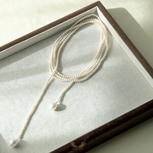 May include: A delicate pearl necklace with a lariat design. The necklace features multiple strands of small, creamy white pearls that cascade down, ending with two larger pearls. The necklace is displayed in a wooden tray with a white linen lining.