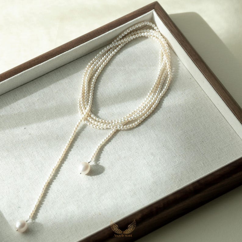 May include: A delicate pearl necklace with a lariat design. The necklace features multiple strands of small, creamy white pearls that cascade down, ending with two larger pearls. The necklace is displayed in a wooden tray with a white linen lining.