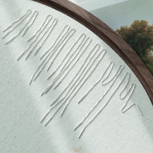 Puede incluir: Primer plano de un bastidor de bordado con hilo blanco cosido sobre tela de color claro. El bastidor de madera enmarca el dise&ntilde;o, que presenta l&iacute;neas verticales y curvas, creando una est&eacute;tica minimalista. El fondo est&aacute; borroso.