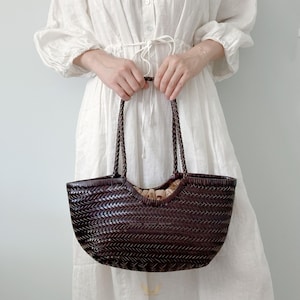 May include: A dark brown woven tote bag with braided handles. The bag has a wide, open top and a rounded shape. The interior appears to be lined with a light-colored fabric. The bag is held by a person wearing a white dress.