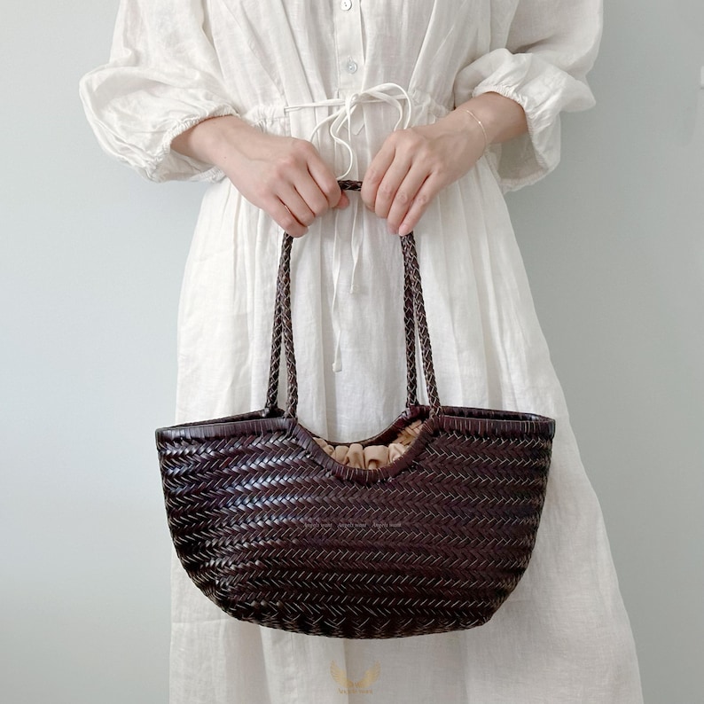 May include: A dark brown woven tote bag with braided handles. The bag has a wide, open top and a rounded shape. The interior appears to be lined with a light-colored fabric. The bag is held by a person wearing a white dress.