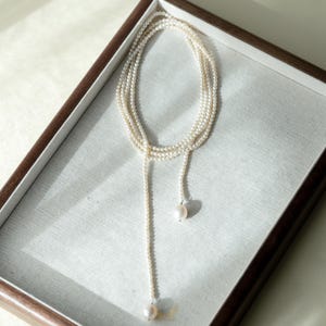 May include: A delicate pearl necklace with a lariat design. The necklace features multiple strands of small, creamy white pearls that cascade down to a single, larger pearl pendant. The necklace is displayed in a wooden-framed tray.