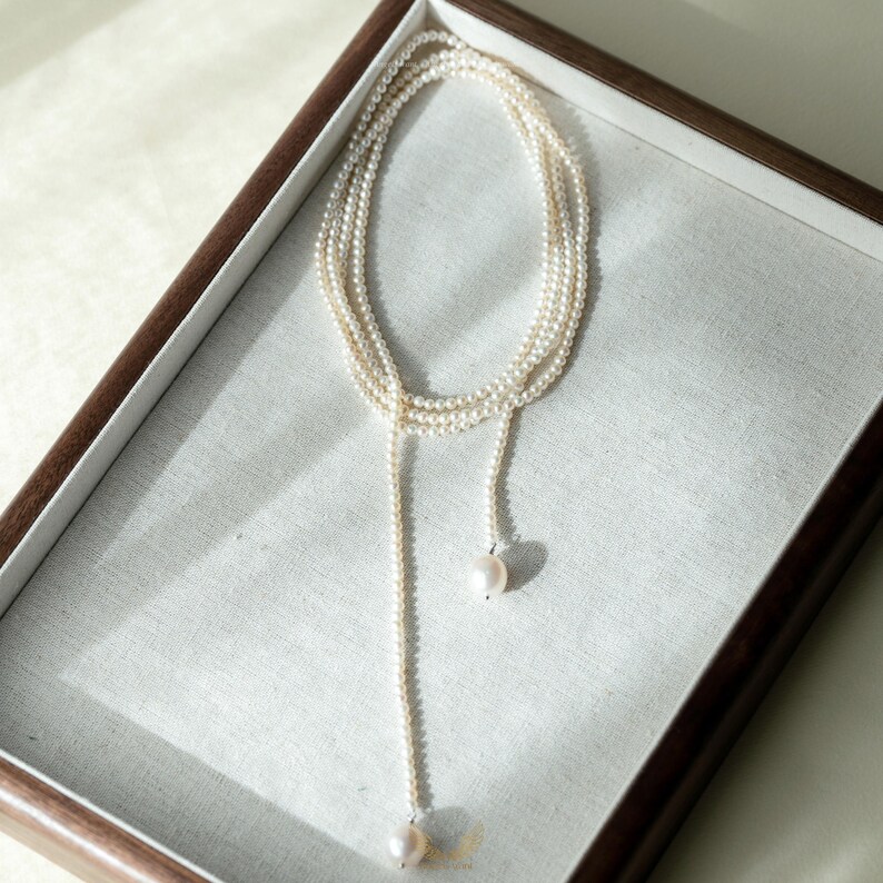 May include: A delicate pearl necklace with a lariat design. The necklace features multiple strands of small, creamy white pearls that cascade down to a single, larger pearl pendant. The necklace is displayed in a wooden-framed tray.