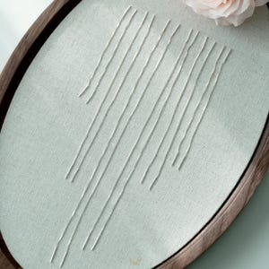 Puede incluir: Un bastidor de bordado ovalado con un fondo de tela verde claro. L&iacute;neas de hilo blanco est&aacute;n cosidas sobre la tela. El bastidor tiene un marco de madera marr&oacute;n oscuro. Una rosa rosa est&aacute; en la esquina superior derecha.