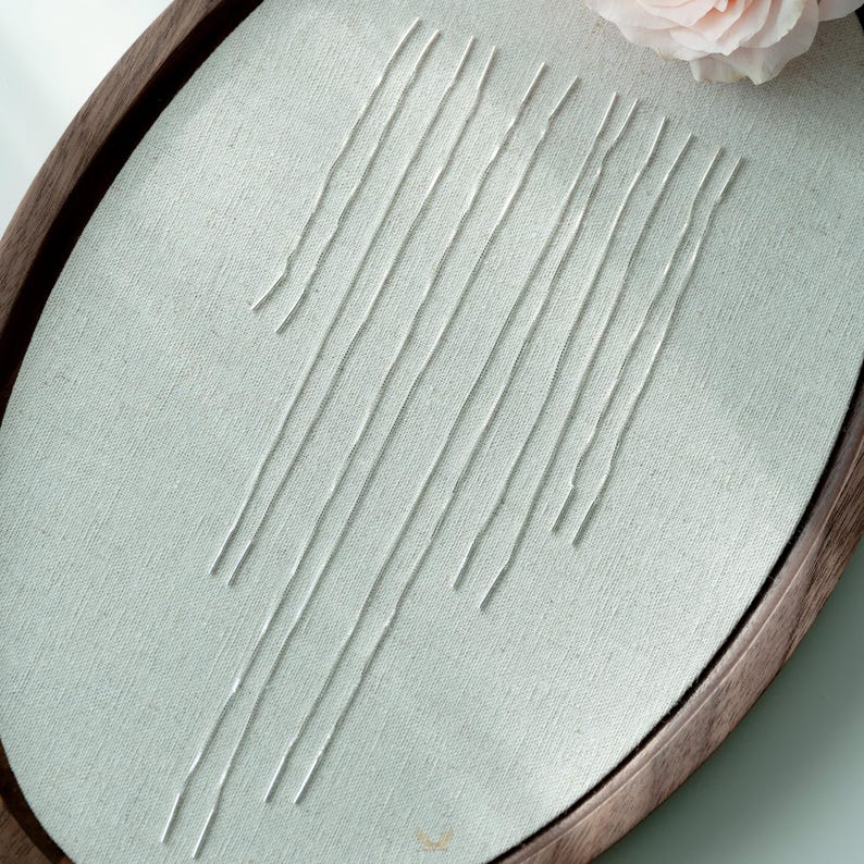 Puede incluir: Un bastidor de bordado ovalado con un fondo de tela verde claro. L&iacute;neas de hilo blanco est&aacute;n cosidas sobre la tela. El bastidor tiene un marco de madera marr&oacute;n oscuro. Una rosa rosa est&aacute; en la esquina superior derecha.