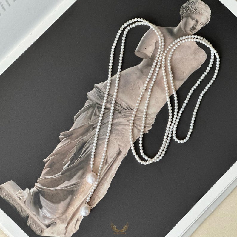 May include: Two long strands of white pearl necklaces draped over a classical statue. The pearls are small and evenly spaced, with a larger pearl at the end of each strand. The statue is a light beige color, set against a dark gray background.