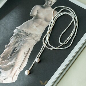 May include: A delicate pearl necklace with a lariat design. The necklace features multiple strands of small, white pearls that cascade down, ending with two larger, round pearls. The jewelry is displayed on a black and white image of a classical statue.