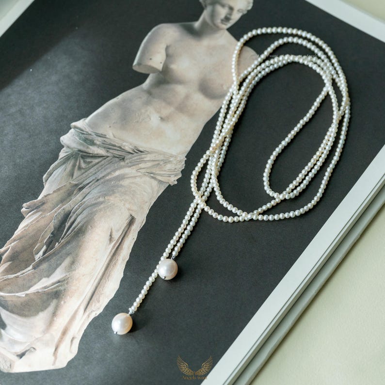 May include: A delicate pearl necklace with a lariat design. The necklace features multiple strands of small, white pearls that cascade down, ending with two larger, round pearls. The jewelry is displayed on a black and white image of a classical statue.