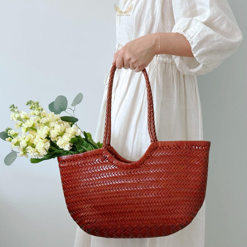 May include: A woven, reddish-brown tote bag with braided handles. The bag is filled with a bouquet of white and yellow flowers and green foliage. The bag is held by a person wearing a white dress.