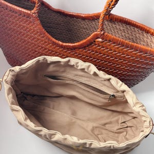 May include: A woven brown leather tote bag with braided handles. The bag has a spacious interior and a tan drawstring pouch with a zippered pocket. The bag is designed for carrying items.