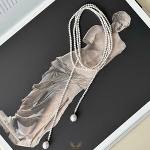 May include: A long, layered pearl necklace with a draped design. The necklace features small, round pearls and larger, single pearls at the ends. The necklace is displayed on a statue against a dark background.