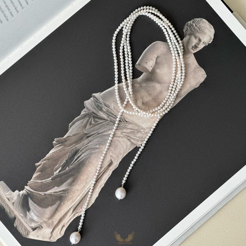 May include: A long, layered pearl necklace with a draped design. The necklace features small, round pearls and larger, single pearls at the ends. The necklace is displayed on a statue against a dark background.