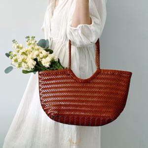 May include: A woven brown leather tote bag with braided handles. The bag is filled with a bouquet of white and yellow flowers with green foliage. The bag has a curved top and a rectangular shape.