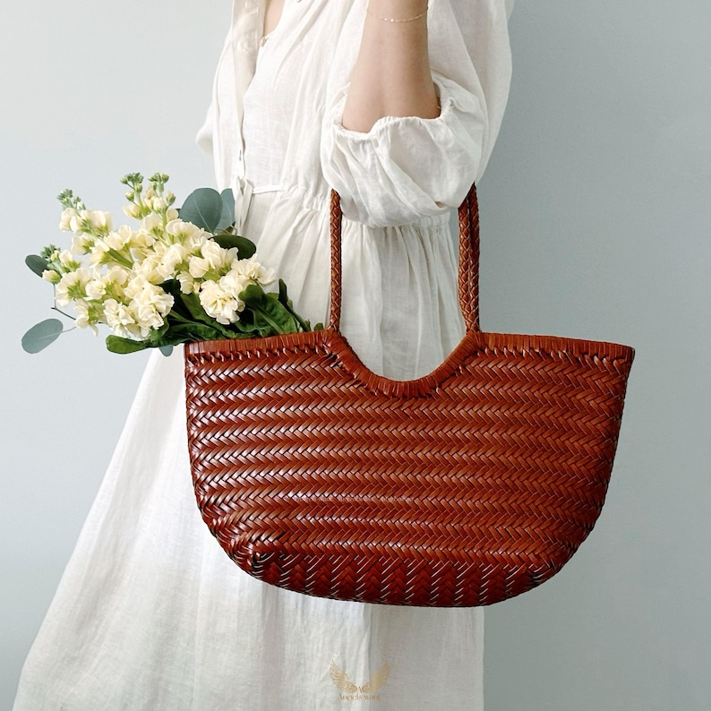 May include: A woven brown leather tote bag with braided handles. The bag is filled with a bouquet of white and yellow flowers with green foliage. The bag has a curved top and a rectangular shape.