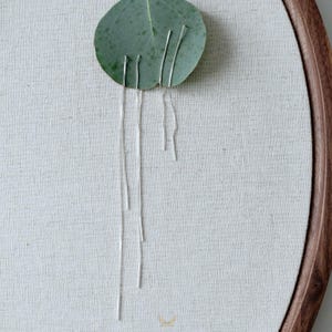 Puede incluir: Primer plano de una pieza decorativa con una hoja verde y cadenas plateadas adjuntas. La hoja est&aacute; fijada a una tela de color claro estirada dentro de un bastidor de bordado de madera. Las cadenas cuelgan, creando un efecto visual &uacute;nico.