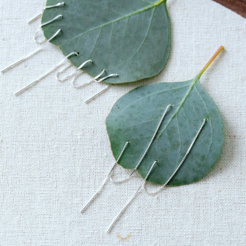 Puede incluir: Pendientes de hilo plateados con un dise&ntilde;o lineal. Los pendientes se muestran sobre una hoja verde, sobre un fondo neutro. Los pendientes tienen una delicada cadena y una barra recta en el extremo.