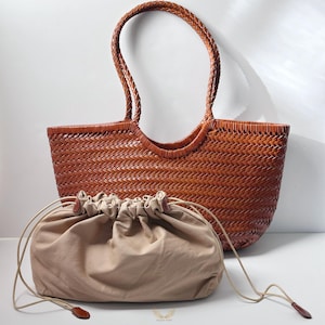 May include: A woven brown leather tote bag with braided handles and a matching drawstring pouch. The tote bag has a rectangular shape with a wide opening. The drawstring pouch is beige and has a leather pull.