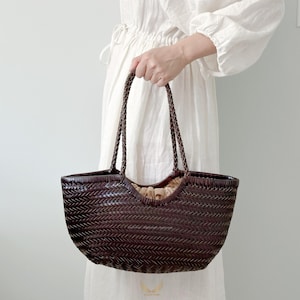 May include: A dark brown woven tote bag with braided handles. The bag has a rounded shape and a light-colored interior. The bag is held by a person wearing a white dress.