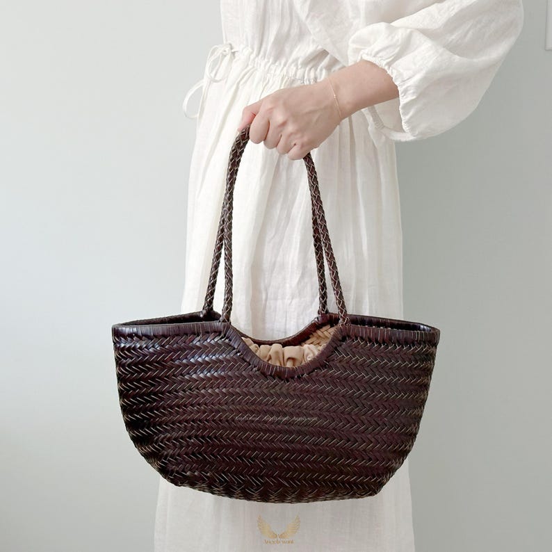 May include: A dark brown woven tote bag with braided handles. The bag has a rounded shape and a light-colored interior. The bag is held by a person wearing a white dress.