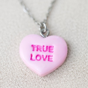 May include: A pink heart-shaped pendant necklace with the words "TRUE LOVE" embossed in a darker pink. The pendant hangs from a silver chain. The necklace is displayed on a textured, light-coloured surface.