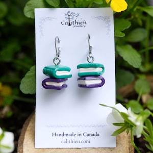 May include: A pair of dangle earrings featuring miniature stacked books in teal and purple with white pages. The earrings are displayed on a white card labeled Calithien Jewellery and Handmade in Canada, featuring silver-toned metal hooks.
